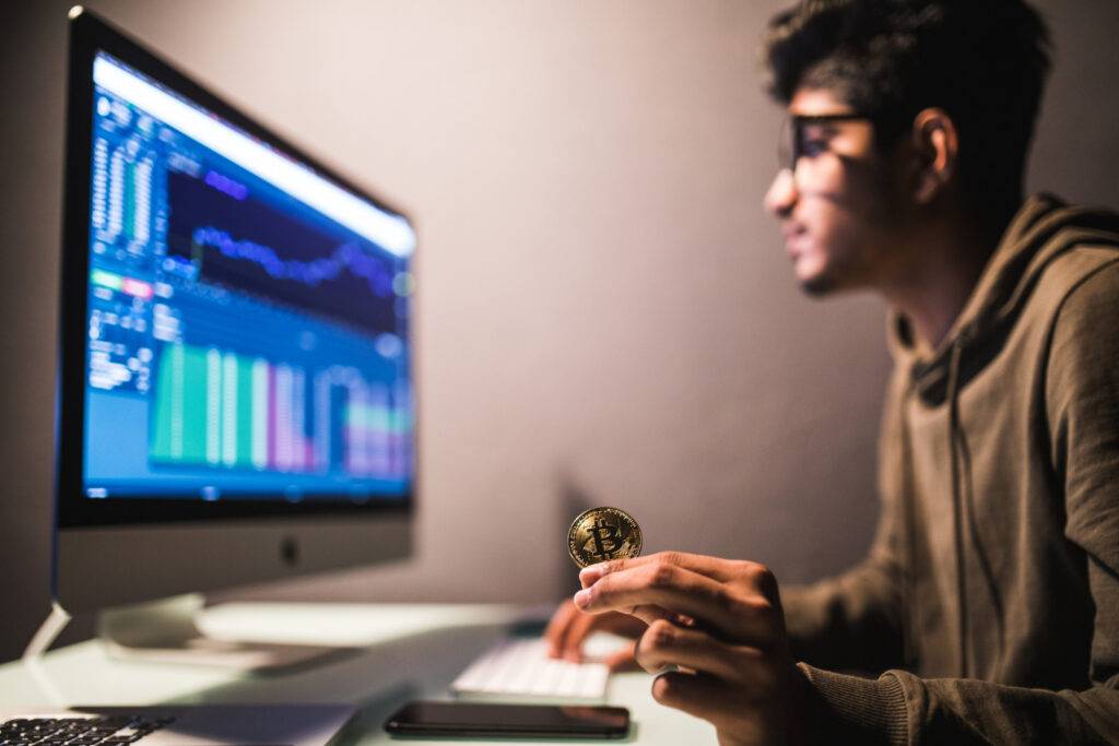 Man analyzing crypto charts on a computer while holding a Bitcoin, relating to a crypto exchange roadmap.